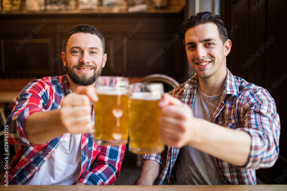 Having a pint with friend. Two cheerful young men toasting with beer while sitting together at the bar