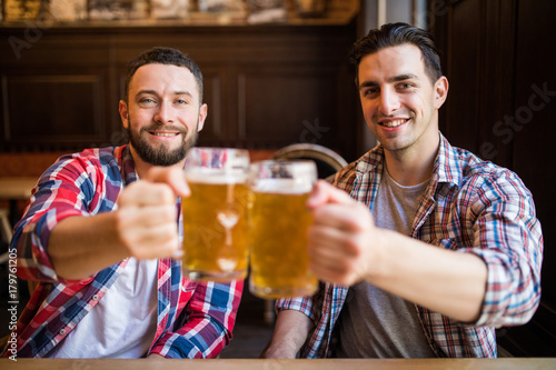 Having a pint with friend. Two cheerful young men toasting with beer while sitting together at the bar