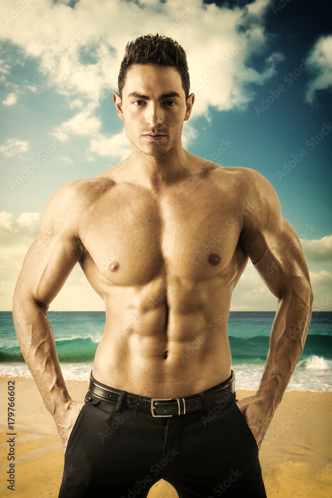 Attractive muscular man by the sea, shirtless looking at camera, standing in the beach