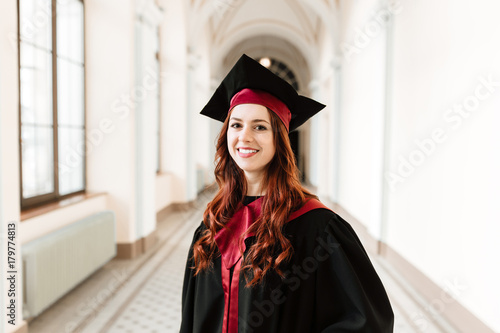 Beautiful, happy, fun, girl graduate in the mantle standing in the hallway of the university and smiles, study concept, space for text