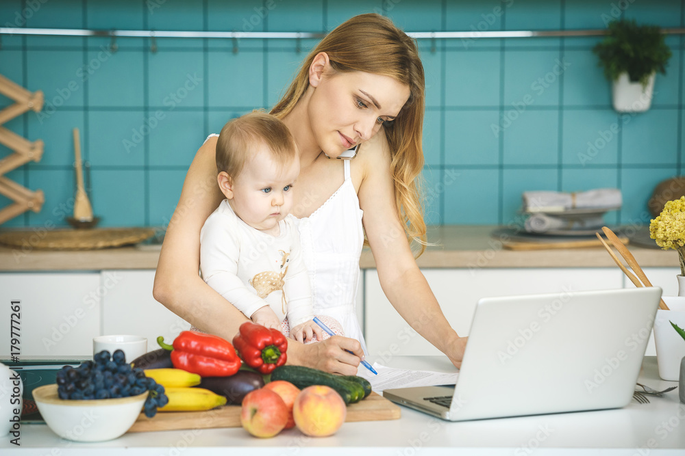 Young mother smiling, cooking and playing with her baby daughter in a modern kitchen. Using phone. Healthy food concept. Working at home. Young mother smiling, cooking and playing with her baby daughter in a modern kitchen. Using phone. Healthy food concept. Working at home.