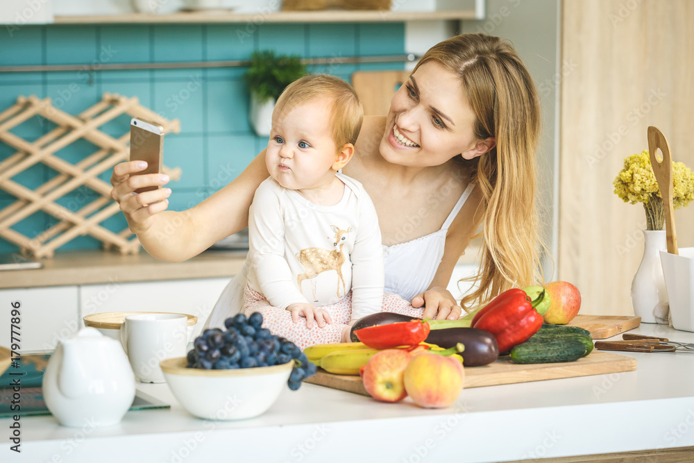 I love selfie! Young mother looking at camera and smiling, cooking and playing with her baby daughter in a modern kitchen setting. Healthy food concept. I love selfie! Young mother looking at camera and smiling, cooking and playing with her baby daughter in a modern kitchen setting. Healthy food concept.