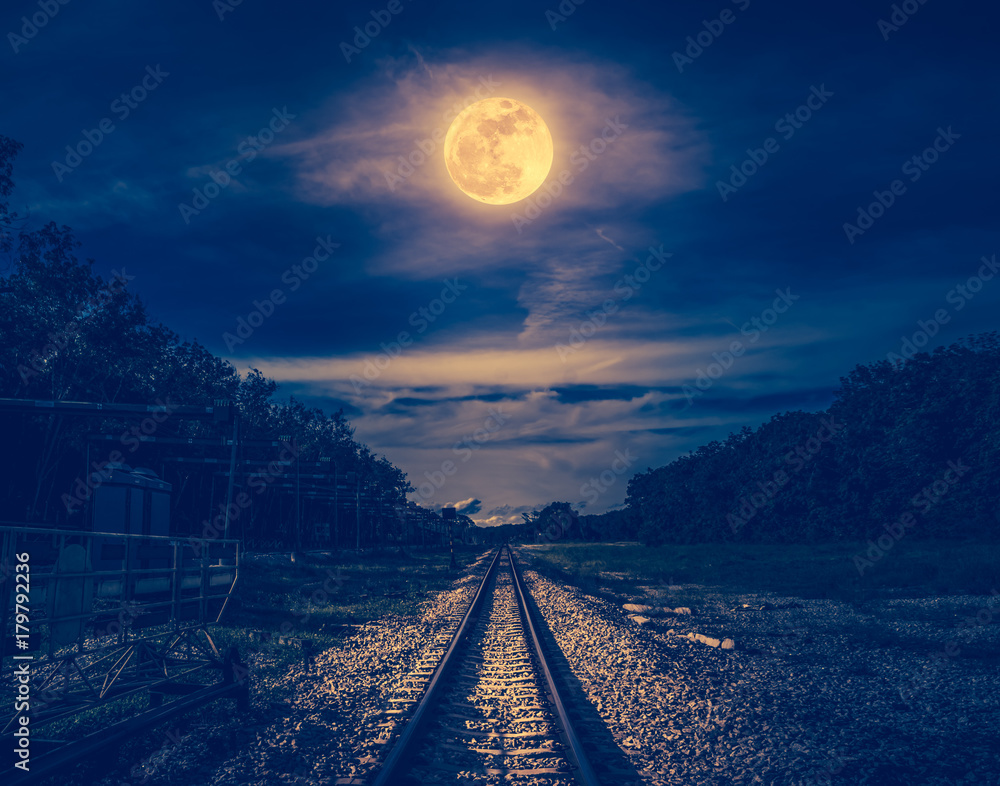 Night sky and full moon above silhouettes of trees and railway.