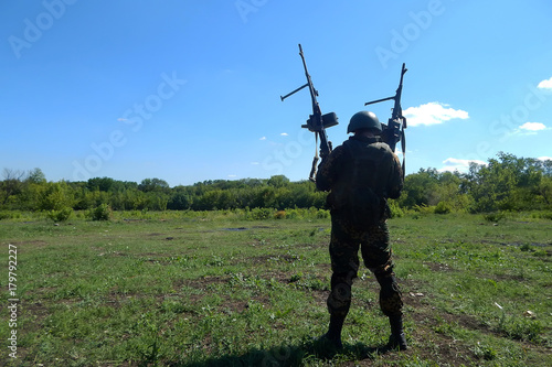 soldier in uniform with two machine guns in his hands is standing on a green field