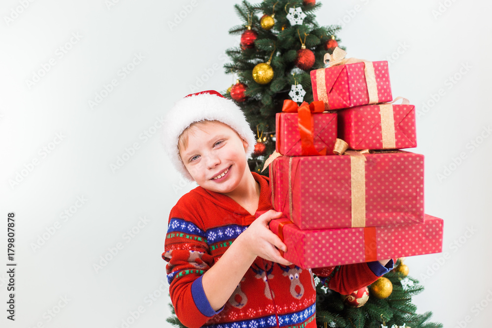 Closeup portrait of happy smiling child holding pile of present boxes in hands ready to open them. Boy wearing santa hat and red pajamas. Holiday green tree in background. Horizontal color photography Closeup portrait of happy smiling child holding pile of present boxes in hands ready to open them. Boy wearing santa hat and red pajamas. Holiday green tree in background. Horizontal color photography