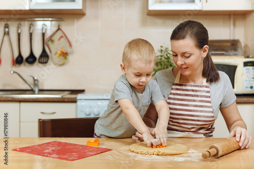 Little kid boy helps mother to cook Christmas ginger biscuit in light kitchen. Happy family mom 30-35 years and child 2-3 roll out dough and cut out cookies at home. Relationship and love concept