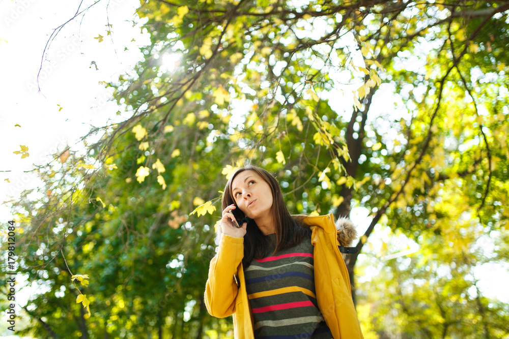 A beautiful happy cheerful brown-haired woman in a yellow coat and striped longsleeve talking on mobile phone in fall city park on a warm day. Autumn golden leaves. A beautiful happy cheerful brown-haired woman in a yellow coat and striped longsleeve talking on mobile phone in fall city park on a warm day. Autumn golden leaves.