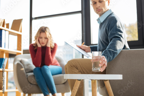 Being thirsty. Sad cheerless depressed man holding his notes and taking a glass of water while having a session with his patient