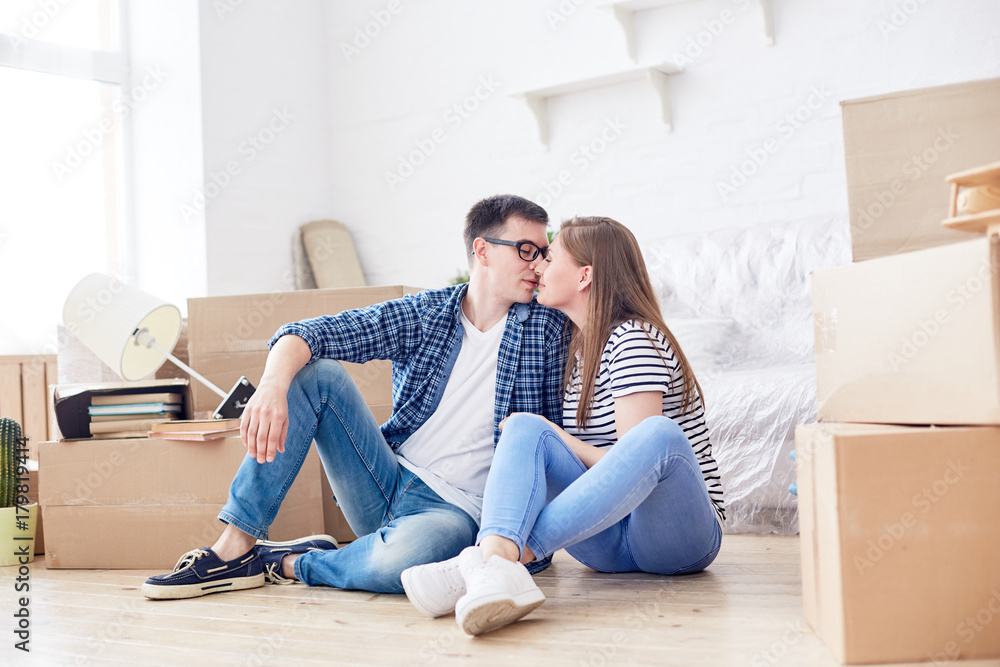 Full length portrait of loving young couple expressing their feeling in kiss while sitting on floor of living room surrounded with moving boxes