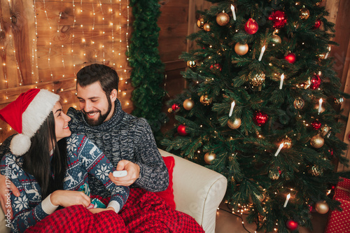 Merry Christmas and Happy New Year!. Young couple celebrating holiday at home. The man is holding the remote from the TV. A guy and a young woman are watching television photo