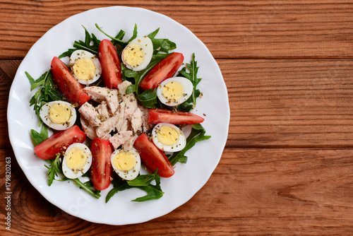 Fresh vegetable chicken salad idea for lunch or dinner. Salad with fresh tomatoes, arugula, quail eggs, chicken fillet and spices on a white plate and a wooden background with copy space. Top view © onlynuta