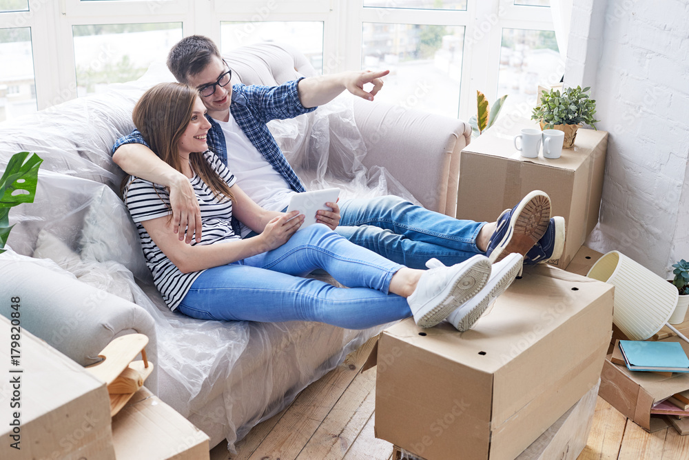 Dreamy young man in eyeglasses embracing his pretty girlfriend while sitting on sofa and sharing ideas concerning interior design of their new apartment, piles of moving boxes Dreamy young man in eyeglasses embracing his pretty girlfriend while sitting on sofa and sharing ideas concerning interior design of their new apartment, piles of moving boxes