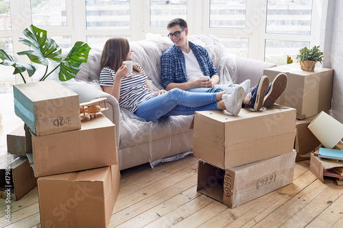 Cheerful young couple chatting animatedly with each other and drinking delicious cappuccino while taking short break from unpacking stuff, piles of moving boxes
