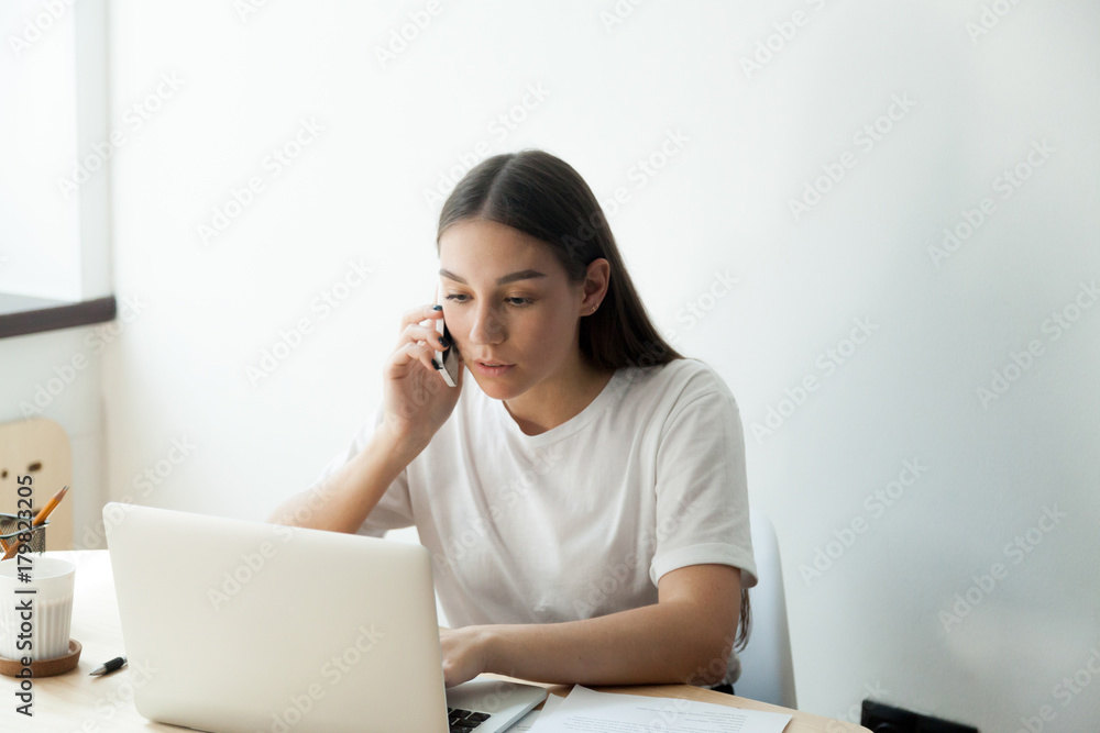 Attentive young adult woman check her order in laptop computer and confirms details by smartphone. Corporate employee planning her vacation. Attentive young adult woman check her order in laptop computer and confirms details by smartphone. Corporate employee planning her vacation.
