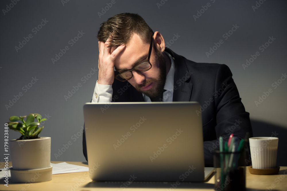 Too much work concept. Troubled man in glasses reading about recent problem on laptop computer, deep in thoughts about best course of actions. Hand supports head, covering forehead, face palm gesture Too much work concept. Troubled man in glasses reading about recent problem on laptop computer, deep in thoughts about best course of actions. Hand supports head, covering forehead, face palm gesture