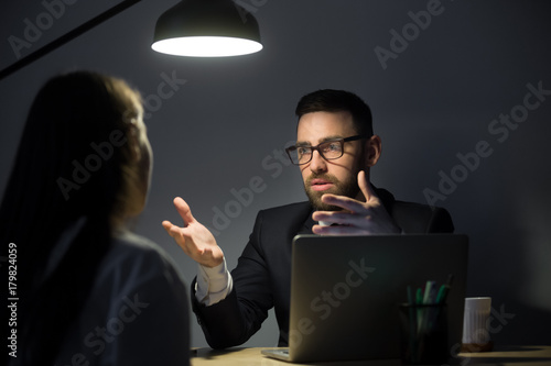 Two businesspeople communicating in late evening meeting in dark office, discussing problem with new project. Man gesturing, persuading business partner.  Colleagues teamwork, late work concept. photo