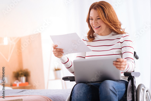 Enjoying work. Beautiful alive blond woman of middle age smiling and holding a sheet of paper and her laptop while sitting in the wheelchair photo