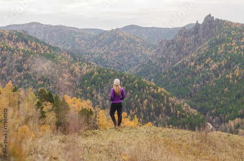 A girl in a lilac jacket looks out into the distance on a mountain, a view of the mountains and an autumnal forest by an overcast day. Free space for text © Maksim