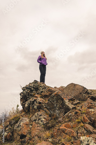A girl in a lilac jacket looks out into the distance on a mountain  a view of the mountains and an autumnal forest by an overcast day  free space for text
