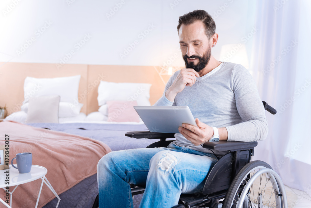 Having a chat. Gleeful smiling bearded disabled man touching his chin and using a tablet while sitting in a wheelchair Having a chat. Gleeful smiling bearded disabled man touching his chin and using a tablet while sitting in a wheelchair