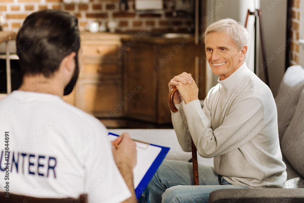Obraz premium Cheerful pensioner. Pleasant positive elderly man looking happy while sitting near his kind attentive volunteer and smiling to him