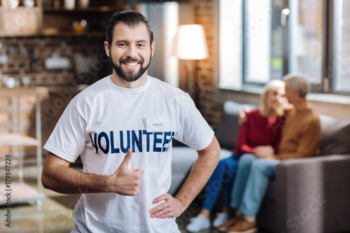 Happy worker. Cheerful positive young volunteer looking glad while helping senior people sitting behind his back