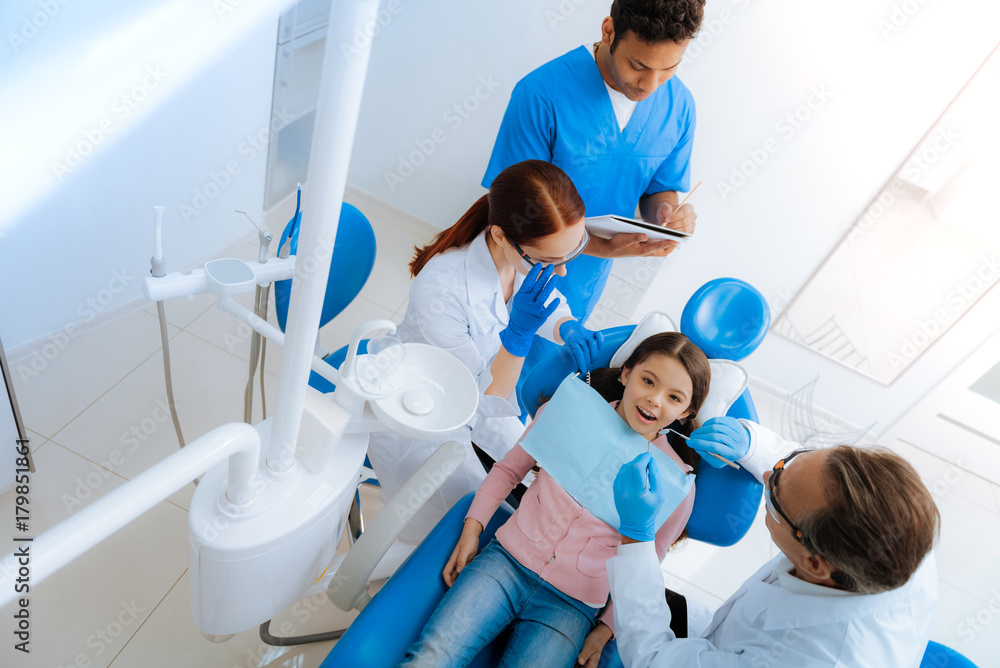 Dentists office. Top view of a nice cute pleasant girl sitting in the dentists chair and opening her mouth while having a medical examination on her teeth Dentists office. Top view of a nice cute pleasant girl sitting in the dentists chair and opening her mouth while having a medical examination on her teeth