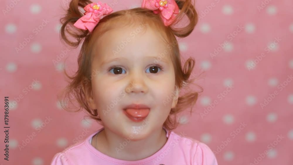 Funny little girl eating a candy with a picture of the heart. A happy cute baby holds a candy in his hand and expresses different emotions - smiling, teasing and showing the tongue. Close-up. Funny little girl eating a candy with a picture of the heart. A happy cute baby holds a candy in his hand and expresses different emotions - smiling, teasing and showing the tongue. Close-up.