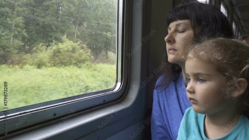Train departs from station. Little curious girl and her mother looking out of window in train. Drops of rain on glass. It's raining outside, people reflecting in glass. Train departs from station. Little curious girl and her mother looking out of window in train. Drops of rain on glass. It's raining outside, people reflecting in glass.