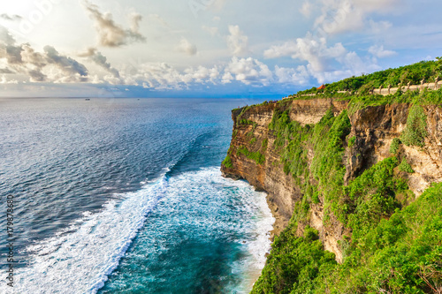 Fantastic huge promontory, rocks are overgrown with green vegetation, stone fence along the cliff, waves far below crash into the rocks. Pura Uluwatu viewpoint, horizontal frame, Bali, Indonesia. photo