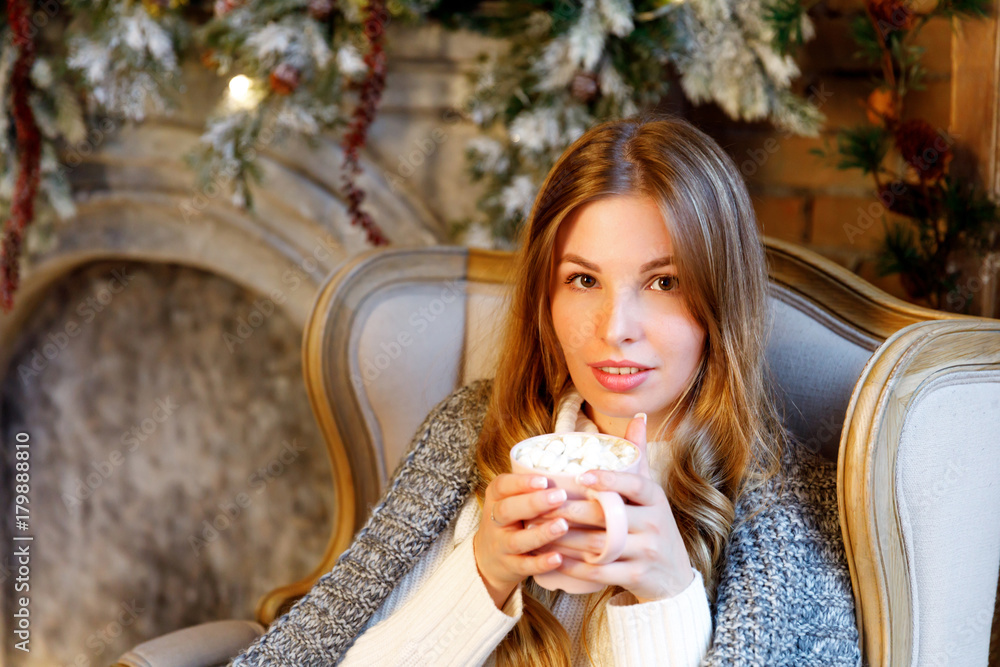Girl in white knitted jumper with cocoa marshmallow mug in the armchair by the fireplace. Winter holidays Girl in white knitted jumper with cocoa marshmallow mug in the armchair by the fireplace. Winter holidays