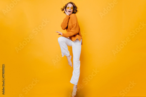 Amazing short-haired woman in trendy sweater standing on one leg and propping face with hand. Indoor full-length portrait of enthusiastic barefooted girl wears white pants.