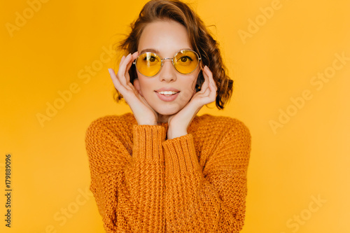Gorgeous white young lady in trendy sweater propping face with hands and looking with interest. Close-up indoor photo of pretty european woman in sunglasses posing on yellow background. © Look!