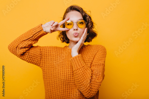 Stunning short-haired female model posing with kiss face expression on yellow background. Close-up portrait of stylish european girl standing with peace sign in front of wall.