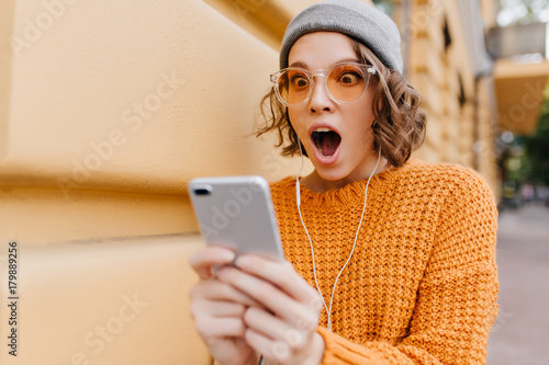 Shocked girl with brown eyes looking at phone screen with mouth open. Outdoor portrait of surprised young woman with curly hair wearing yellow knitted attire and holding smartphone. photo