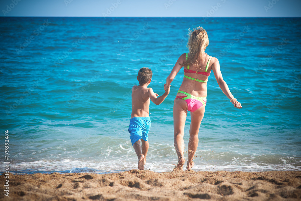 Caucasian mom and son running into the sea water. Back view. Caucasian mom and son running into the sea water. Back view.