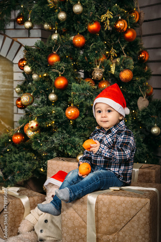 Christmas portrait of happy smiling little boy in red santa hat sitting on boxes with presents holding oranges in hands near the christmas tree. Winter holiday Xmas and New Year concept