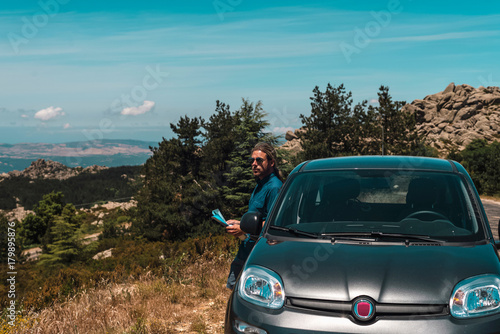 Male tourist leaning against car parked in mountains. Sardinia. Italy.