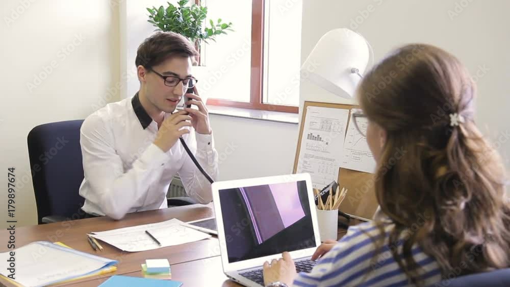 Male employee with telephone specifies information from woman opposite. Man with glasses asking a question at an employee in office behind a desk. Male employee with telephone specifies information from woman opposite. Man with glasses asking a question at an employee in office behind a desk.