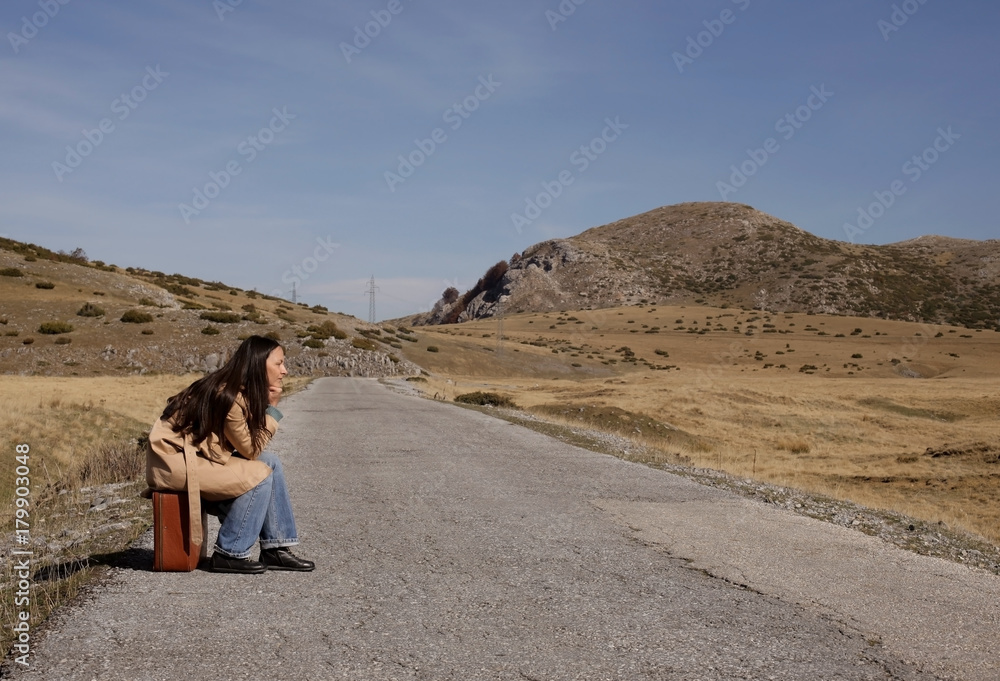 Woman traveler is sitting on her suitcase and is waiting someone to come; woman traveler on an empty road  on mountain; people and travel concept.