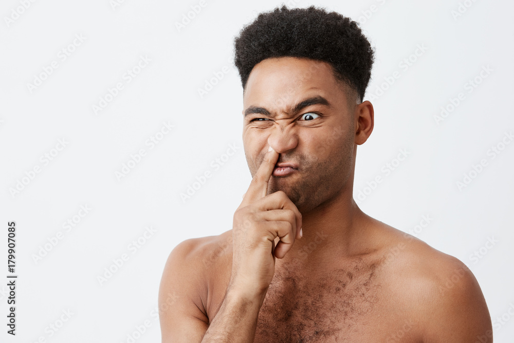 Close up of silly beautiful dark-skinned american with afro hairstyle and naked body holding finger in nose, looking aside with stupid face expression, being on his un in morning.