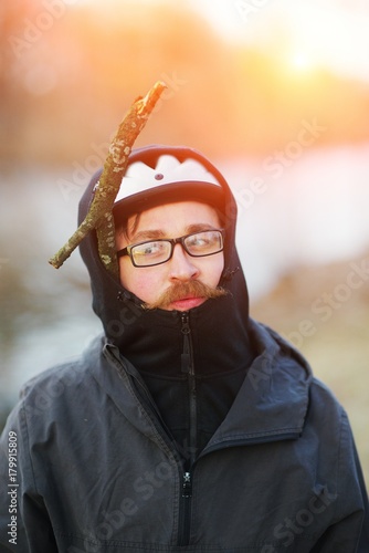 Artistic portrait young pranksters of man with the big red mustache with glasses and a bike helmet and with dry branch under bonnet on blurred background of Park. Close-up photo