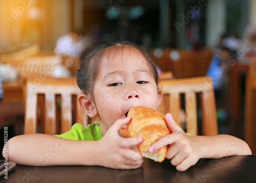 Asian little girl eating croissant at restaurant.