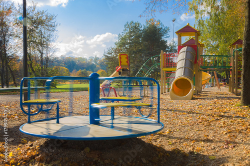 Children playground consisting of rotating carousel, swing, tube and climbing frame. Fallen autumn leaves of trees on the background