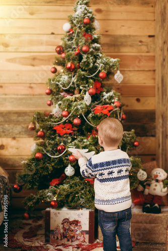Happy cute little baby boy dressed in sweater and jeans decorating Christmas tree with toys in wooden room at home. Child with good mood. New Year. Lifestyle, family and holiday 2018 concept