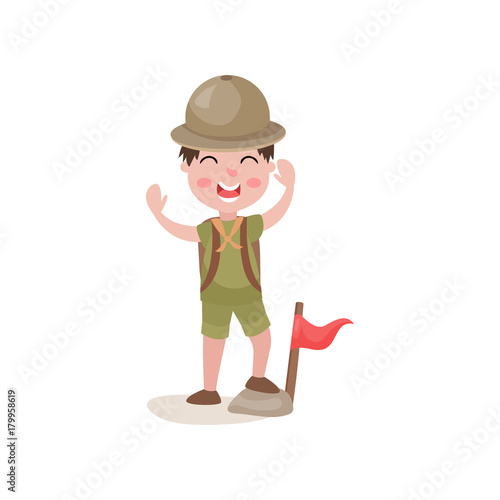 Boy scout standing with one foot on stone near flag and cheerfully waving his hands