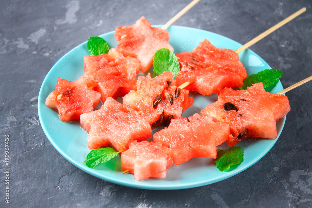 Watermelon in the form of stars on skewers with leaves of mint lies on a plate. The blue dish is like a rocket in space. Top view. Watermelon in the form of stars on skewers with leaves of mint lies on a plate. The blue dish is like a rocket in space. Top view.