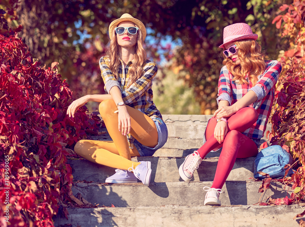 Fall Fashion. Urban Outdoor. Young Woman Sitting on Steps, Relax Enjoy  Nature. Fashion. Sisters Best Friends in Stylish Autumn Hipster Outfit,  Sunglasses. Model Girl in Sunny Colorful Park foto de Stock
