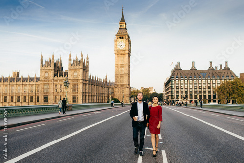 Female wears red elegant dress and man hold hands, have walk on Westmisnter bridge, admire London`s sights, have excursion, like travelling together. Young enthusiastic tourists in Great Britain