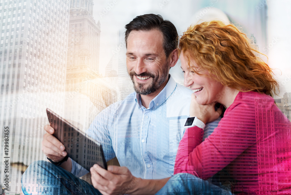Fototapeta premium Amusing content. Pleasant young man and his charming wife watching funny videos on tablet and laughing
